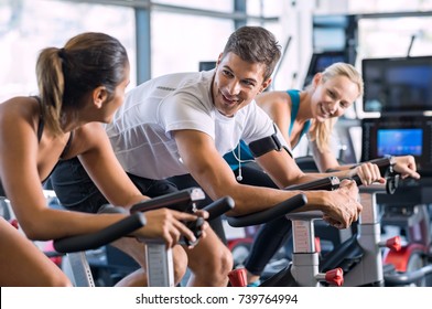 Young People Talking And Smiling While Working Out On Bike At Gym. Friends In A Conversation While Cycling On Stationary Bike In Fitness Centre. Group Of Happy People Working Out At Spinning Class.