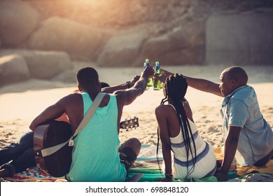 Young people toasting with beer bottles while sitting on beach. Group of friends having drinks together.