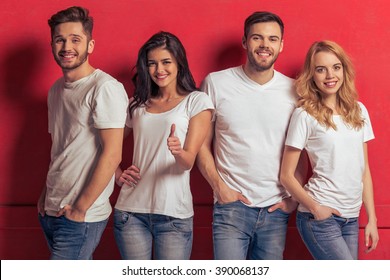 Young People In White T Shirts And Jeans Are Looking At Camera And Smiling, Standing Against Red Background. Girl Is Showing Ok Sign