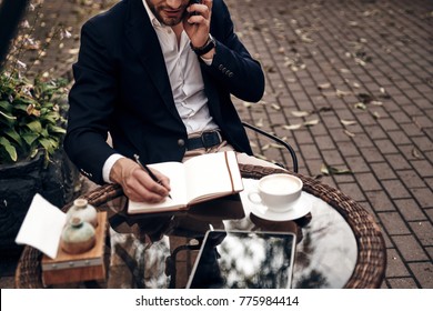 Young Perfectionist. Close Up Top View Of Young Man In Smart Casual Wear Talking On His Smart Phone And Writing Something Down While Sitting In Restaurant Outdoors