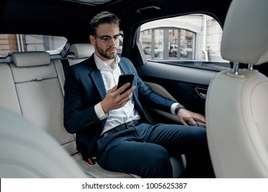 Young Perfectionist. Handsome Young Man In Full Suit Looking At His Smart Phone While Sitting In The Car