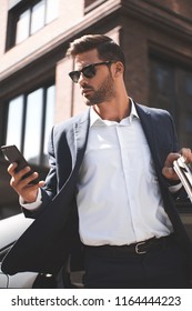 Young Perfectionist. Handsome Young Man In Full Suit Looking At His Smart Phone Near Car