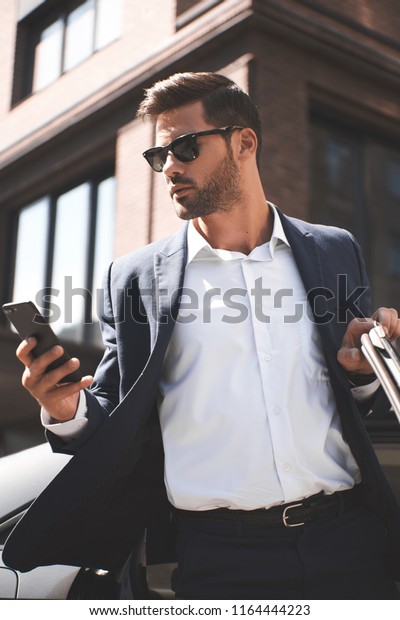 Young perfectionist. Handsome young man in\
full suit looking at his smart phone near\
car