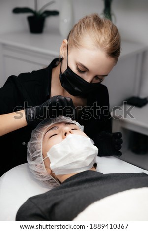 Young permanent makeup artist removes anesthetic cream from the client's eyebrows. Master and client in medical masks and gloves. Permanent makeup eyebrows conturing. New reality in beauty world
