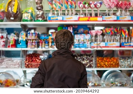 A young person with brown hair wearing a brown casual jacket seen from behind is standing in front of colourful shelves filled with sweets and candy in a store.