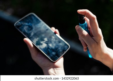 Young Person Cleaning A Mobile Phone Display By Spraying Washing Solution On It Outdoor – Woman Holding A Smartphone While Removing Dirt From The Surface Of The Screen