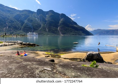 Young Person Jumps Off A Shore Side Diving Board Into The Calm Waters Of A Spectacular Norwegian Fjord, With Children Sunbathing On A Rocky Beach, Eidfjord, Hardangerfjord, Hordaland, Norway 07.12.19