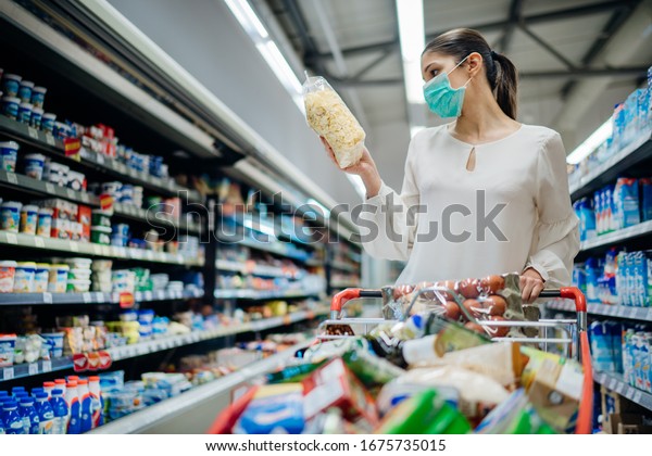 Young person with protective face mask buying\
groceries/supplies in the supermarket.Preparation for a pandemic\
quarantine due to coronavirus covid-19 outbreak.Choosing\
nonperishable food\
essentials