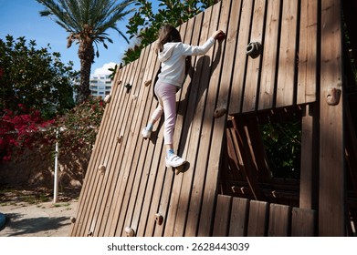 A young person scales a vertical wooden climbing wall with artificial holds in a sunny outdoor playground surrounded by lush greenery and palm trees, enjoying a physically engaging challenge.