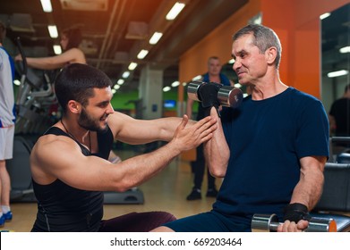 Young Personal Trainer Working With Senior Man Client In Gym. Elderly Man Exercising With Dumbbell. Healthy Lifestyle, Fitness And Sports Concept.