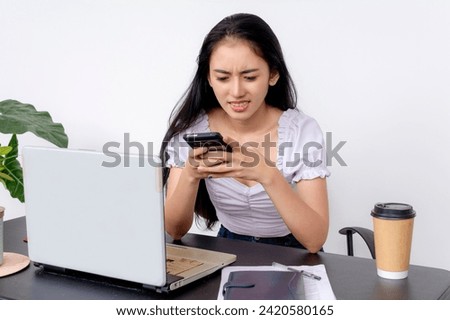 A young perturbed female college student looks stressed as she checks her smartphone at her workspace with a laptop.