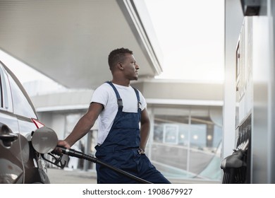 Young Petrol Station Worker, Handsome African Bearded Guy In Workwear, Refueling Luxury Car And Looking Away, While Standing On Gas Station Background