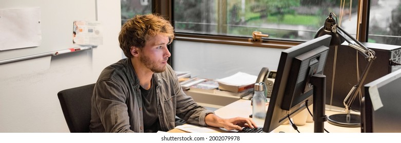 Young PhD student studying writing final paper for exam at university. Working man at office desk typing at desktop computer in office.