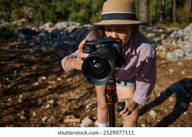 A young photographer adjusts her camera on a tripod in a sunlit outdoor setting, focusing on capturing the perfect shot. Her casual attire and natural surroundings reflect creativity. - Powered by Shutterstock