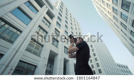 Young photographer takes picture of modern urban architecture. Action. Bottom view of guy photographing modern architecture with camera. Photographer shoots office buildings against blue sky