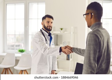 Young Physician Shaking Hands With African-American Man. Doctor Welcoming Client In His Office At The Clinic Or Hospital. Happy Medical Specialist Greeting Patient Before Consultation Or Interview