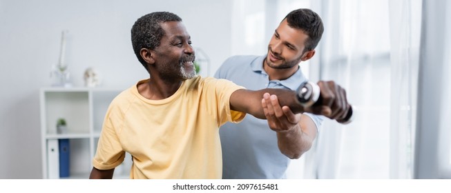 young physiotherapist assisting smiling african american patient exercising with dumbbell, banner - Shutterstock ID 2097615481