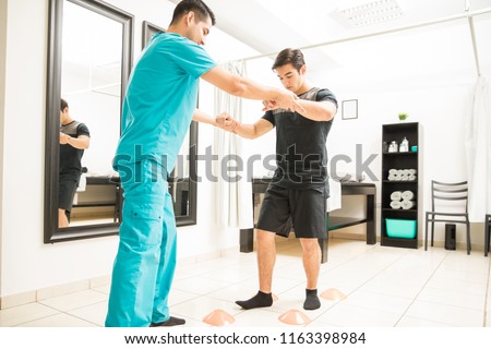 Young physiotherapist helping athlete to walk between cones in hospital