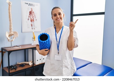 Young Physiotherapist Woman Holding Foam Roll At The Clinic Smiling Happy Pointing With Hand And Finger To The Side 