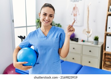 Young physiotherapist woman holding pilates ball at medical clinic smiling happy pointing with hand and finger to the side  - Shutterstock ID 2169823919