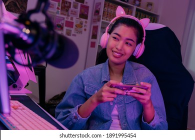 Young playful Asian woman sitting in front of her computer workstation and smiling at the screen with a microphone in foreground. Beautiful asian woman working from her home studio and playing game.