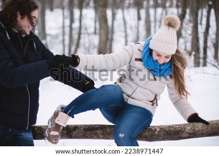 Young playful couple in love outdoors in the forest during winter. The man helps the woman to sit on the barrier.