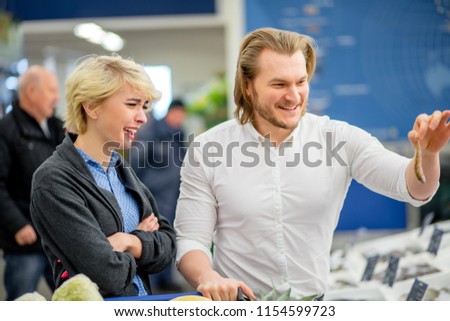 Young playful husband fooling around in fish department, amusing his blonde wife, cheerful couple choosing fish in supermarket.