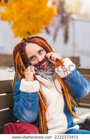 Young playful woman with bright hairstyle in warm clothes on walk in park. Positive female making mustache from dreadlocks, laughing, having fun