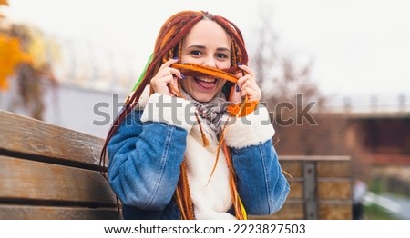 Young playful woman with bright hairstyle in warm clothes on walk in park. Positive female making mustache from dreadlocks, laughing, having fun