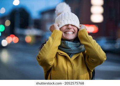 Young playful woman covering eyes with winter cap on city street. Laughing woman on road covering eyes and having fun in a cold winter evening. Happy casual girl having fun during dusk.