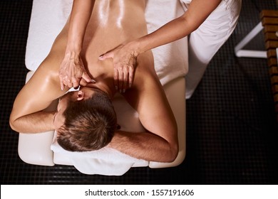 Young Pleasant Man Having Deep Tissue Massage On White Soft Towels, Lying On Black Floor, Gentle Female Hands Massaging Neck Of Male Client, Osteopathy Treatment