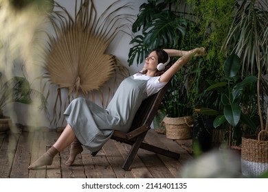 Young Pleased Dreamy Italian Woman With Closed Eyes Wearing Headphones Listening Music To Reduce Stress At Home, Meditating In Stress-relieving Nature Plant Corner At Home. Gardening And Mental Health