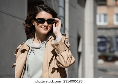young and pleased woman with wavy brunette hair, in grey hoodie and beige trench coat adjusting dark stylish sunglasses and looking away on urban street on blurred background - Powered by Shutterstock