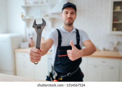 Young Plumber Hold Wrench In Hand. He Show It To Camera And Hold Big Thumb Up. Guy Stand In Kitchen. White Background. Daylight.