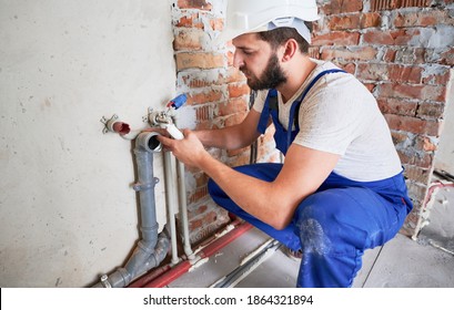 Young Plumber, Wearing Blue Uniform And White Helmet Working With Sealant Fix Of Sewer Pipe In Kitchen Or In Bathroom In Unfinished Apartment. Concept Of Plumbing Works And Home Renovation