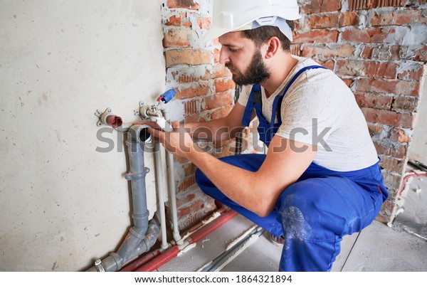 Young plumber, wearing blue uniform and white\
helmet working with sealant fix of sewer pipe in kitchen or in\
bathroom in unfinished apartment. Concept of plumbing works and\
home renovation