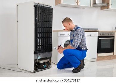 Young Plumber Writing On Clipboard In Front Of Refrigerator Appliance In Kitchen Room