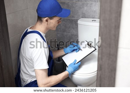Young plumber writing results of examining toilet bowl in water closet