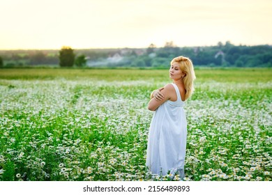 A young plump beautiful woman is resting on a chamomile field at sunset. A plus-size woman in a white sundress in a meadow with daisies