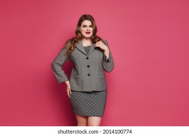 A Young Plump Woman With Bright Makeup And Red Full Lips Wearing Office Clothes Posing On The Pink Background. Plus-size Model Girl In Grey Dress And Suit Coat Over Pink Background