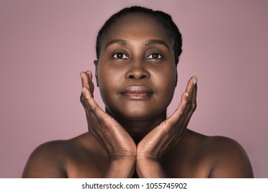 Young plus size African woman with a perfect complexion standing with her hands framing her face against an ash rose colored background