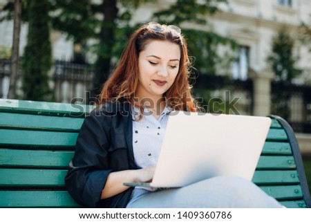 Young  plus size caucasian female with red hair looking at her laptop smiling while sitting on a bench against a building.