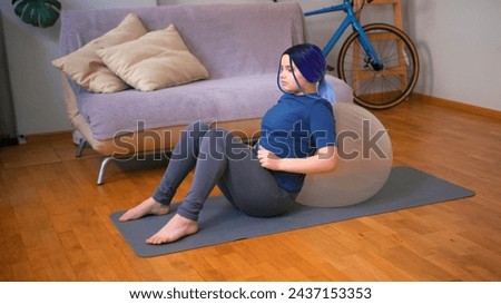 Young plus size latin hispanic teenager girl doing sports at home with fit ball. Cozy home interior with indoor plants. Online fitness, urban jungle concept.