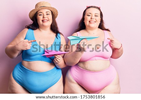 Young plus size twins wearing bikini holding paper airplane smiling happy pointing with hand and finger 