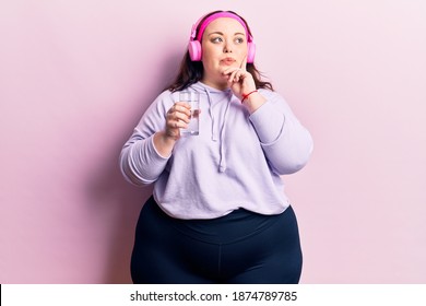 Young plus size woman drinking glass of water using headphones serious face thinking about question with hand on chin, thoughtful about confusing idea 