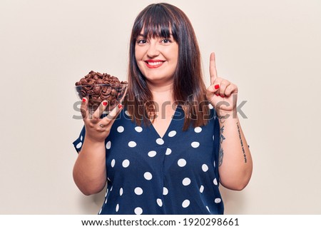 Young plus size woman holding chocolate cereals smiling with an idea or question pointing finger with happy face, number one 