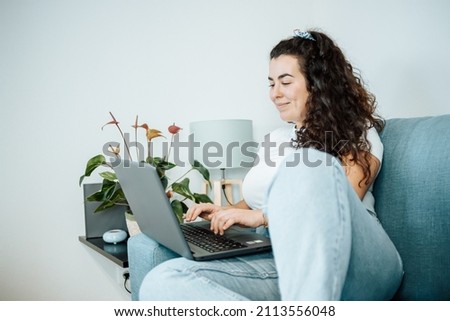 Young plus size woman smiling while working from home on his laptop on the sofa. Young entrepeneur freelancer. Remote tasking work, happy attitude, new job. Modern and young style.