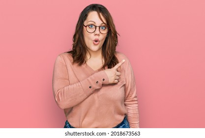 Young plus size woman wearing casual clothes and glasses surprised pointing with finger to the side, open mouth amazed expression. 