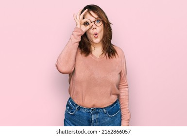 Young plus size woman wearing casual clothes and glasses doing ok gesture shocked with surprised face, eye looking through fingers. unbelieving expression. 