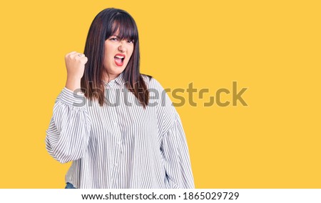 Young plus size woman wearing casual clothes angry and mad raising fist frustrated and furious while shouting with anger. rage and aggressive concept. 